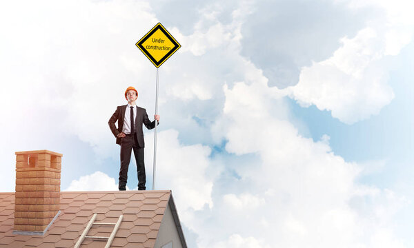 Young engineer on house brick roof holding safety sign indicating under construction notice. Mixed media