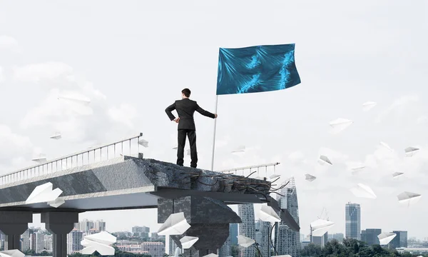 リーダーシップの概念の背景に街並みと壊れた橋に紙飛行機を飛行中に立っている間手に旗を持ってスーツに自信を持っているビジネスマンの背面図 — ストック写真