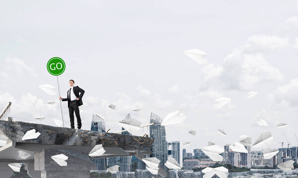 Confident businessman in suit holding green go sign while standing among flying paper planes on broken bridge with cityscape on background. 3D rendering.