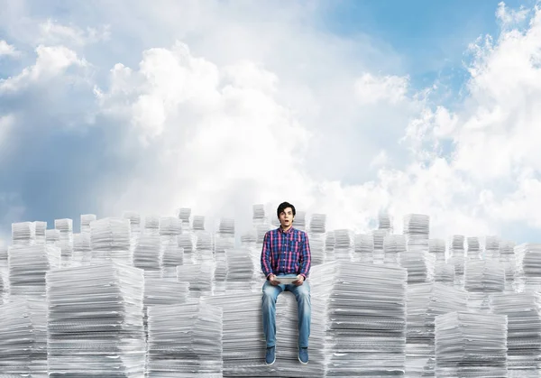 カジュアルウェアの背景に Cloudly 天空と書類の山に座っている若い男 ミクスト メディア — ストック写真