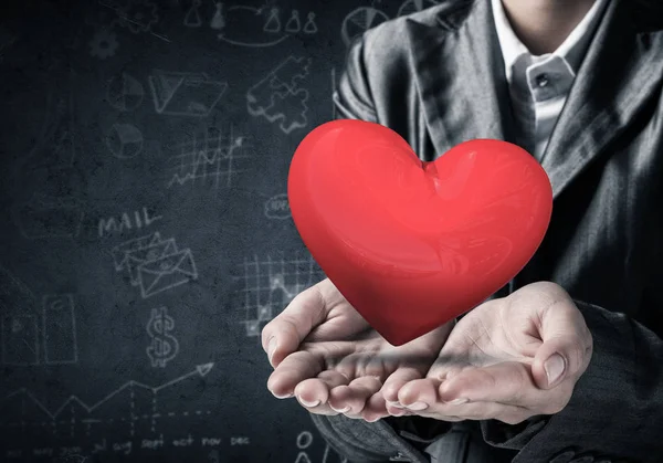 Kırpılmış görüntü arka plan üzerinde koyu duvar skeçler ile elleri büyük kırmızı kalp sunan takım elbiseli işadamı. 3D render.