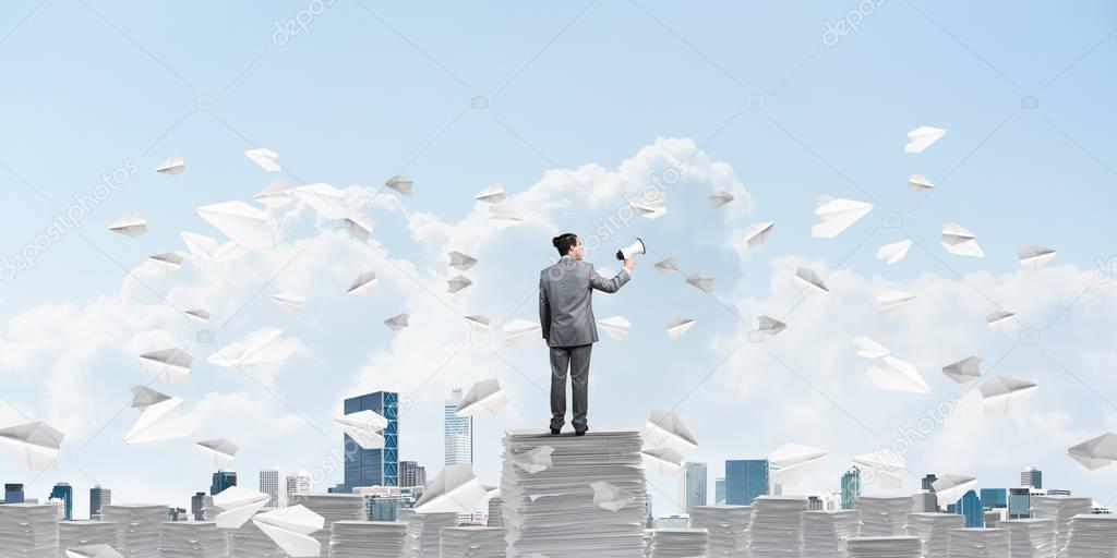 Businessman in suit standing among flying paper planes with speaker in hand and with skyscape on background. Mixed media.