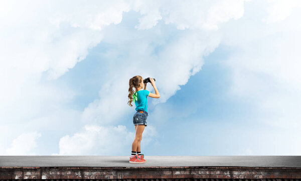 Cute girl of school age on building roof looking in binoculars