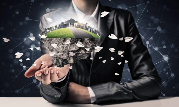 Businessman in suit keeping green island with skycraper city and flying paper planes in hands with network connections on background.