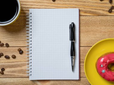 Açılmış not defteri ve bir fincan kahveyle ahşap masada çörek.