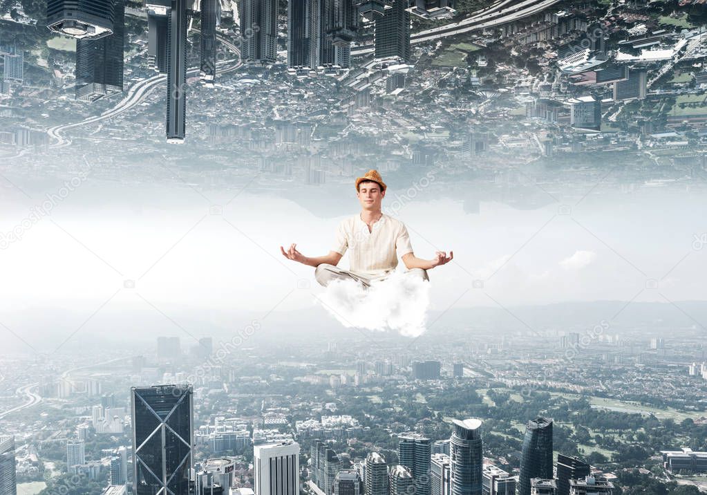 Young man keeping eyes closed and looking concentrated while meditating on cloud in the air between two urban worlds.