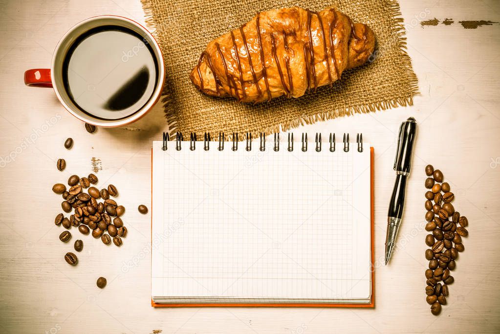 Opened notepad and cup of coffee with croissant on wooden table
