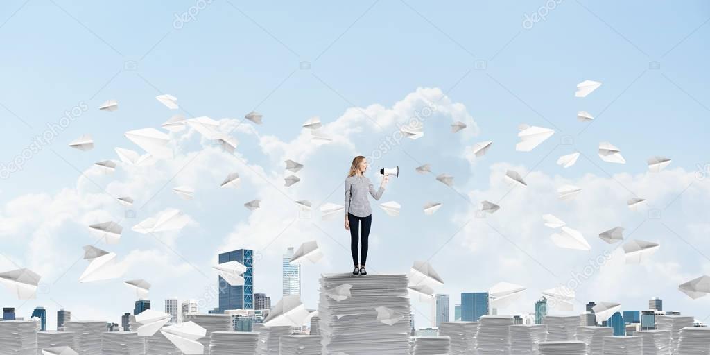 Woman in casual clothing standing among flying paper planes with speaker in hand and with skyscape on background. Mixed media.