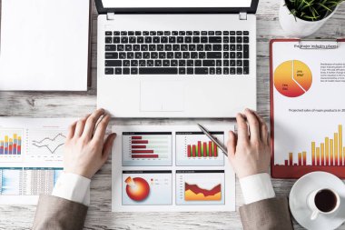 Finansal raporlarla çalışan bir iş adamı. Erkek eli, dizüstü bilgisayarı ve kağıt dökümanları ahşap masanın üzerinde duran düz bir işyeri. Diyagramlı iş sunumu. Yatırım ve borsa analizi.