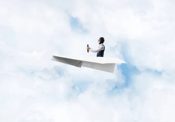 航空機で楽しい飛行士 曇り青空で紙飛行機を運転革のヘルメットとゴーグルのパイロット 小さな紙飛行機で飛ぶ面白い男 ふわふわの雲と雲景の背景 — ストック写真