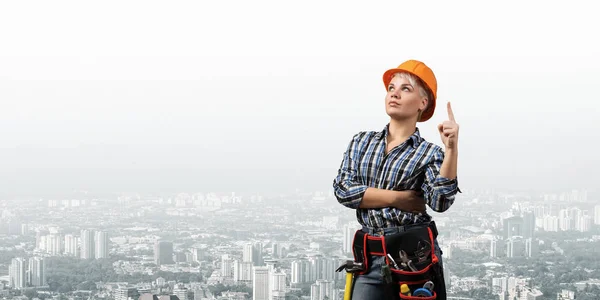漂亮的女建筑工人戴着硬礼帽 手指向上指向上方 穿着格子蓝色衬衫的年轻女子站在城市景观的背景下的画像 工业建筑和建筑 — 图库照片