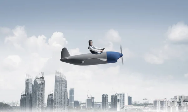 Pilot sitting in cabin of small airplane and holding steering wheel. Funny man in aviator hat and goggles driving propeller plane above city. Modern metropolis with high buildings and towers