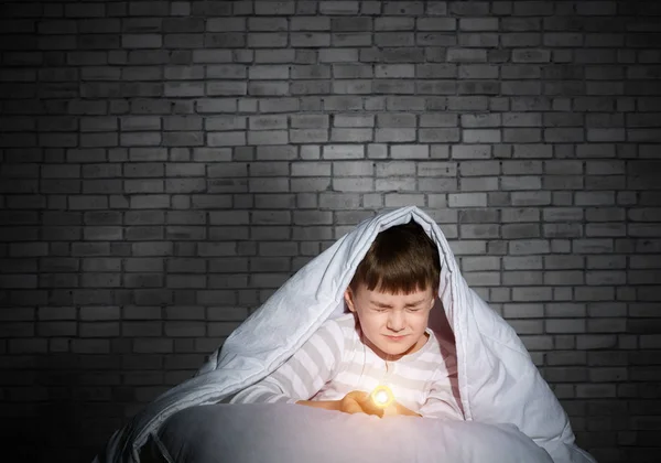 毛布の下に懐中電灯が隠れているおびえた子供 目を閉じた子供が家でベッドに横たわっている怖い子供 小さな男の子は夜眠れない レンガの壁を背景にパジャマ姿の少年の肖像 — ストック写真
