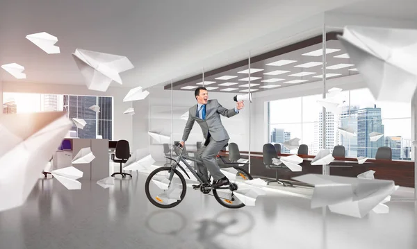 Man in business suit riding bicycle at conference hall. Businessman with megaphone looking back on bike at loft office interior with flying paper planes. Business presentation and announcement.