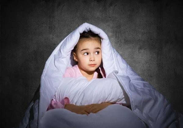 Frightened girl hiding under blanket. Scared kid lying in his bed at home. Fear of the dark. Beautiful little girl can not sleep at night. Portrait of child in pajamas on background of grey wall.