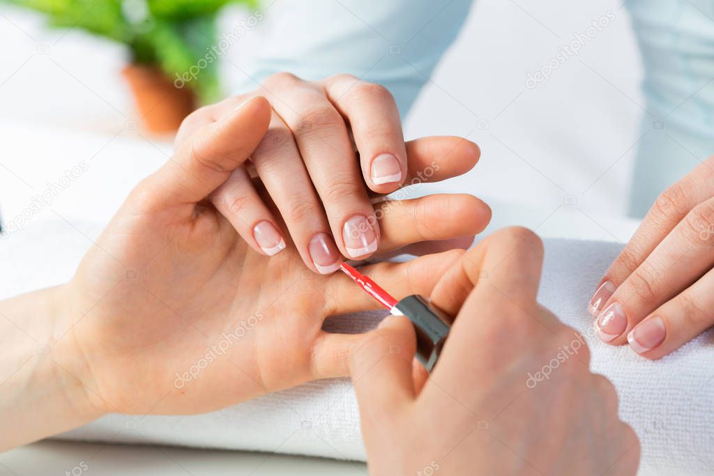 Manicurist working with client's nails at table. Closeup of female hand applying red nail polish in beauty salon. Woman getting nail manicure. Professional nail care and beautician procedure.