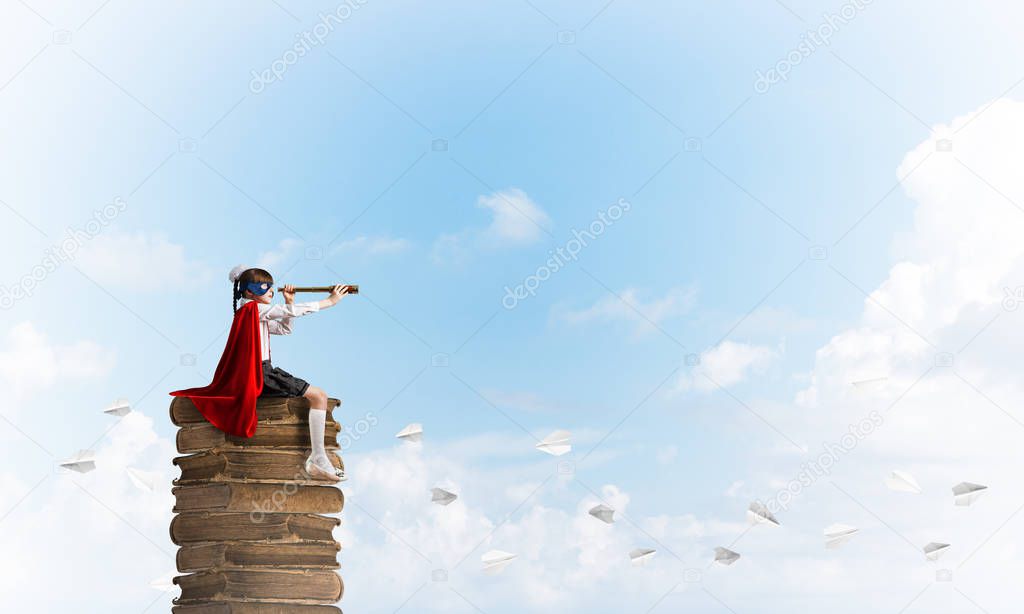 Little child in mask and cape floating on pile of books and looking in spyglass