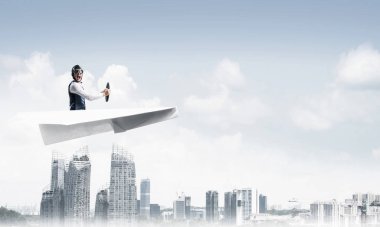 Havacı şapkalı işadamı ve gözlüklü, şehir merkezinin üzerinde yüksek binalarla kağıt uçak sürüyor. Mavi gökyüzü ile modern megalopolis panorama. Karışık medya iş kavramı. Hayal ve hayal gücü.