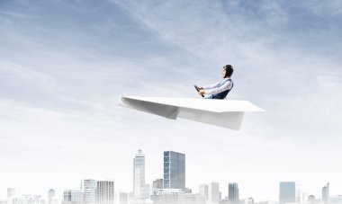 Havacı şapkalı işadamı ve gözlüklü, şehir merkezinin üzerinde yüksek binalarla kağıt uçak sürüyor. Mavi gökyüzü ile modern megalopolis panorama. Karışık medya iş kavramı. Hayal ve hayal gücü.