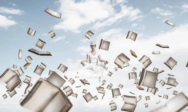 Woman in white clothing keeping eyes closed and looking concentrated while meditating among flying books in the air with cloudy skyscape on background.
