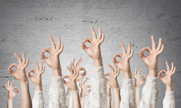 Row of man hands in white sweater showing okay gesture. Agreement and approval group of signs. Human hands gesturing on background of grey wall. Many arms raised together and present popular gesture.