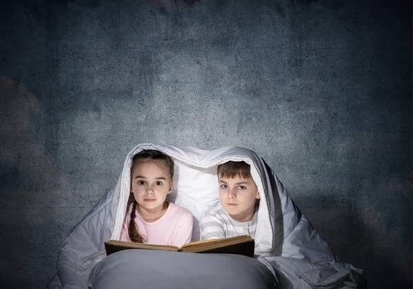 Little girl and boy looking at each other under blanket. Children reading magic stories in bed before going to sleep. Young sister and brother in pajamas together on background of grey wall.