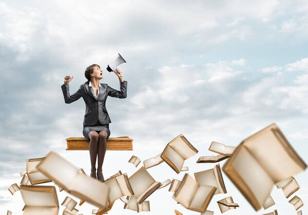 Молодая женщина с мегафоном сидит на большой книге и летает в голубом небе. Красивая деловая женщина на фоне облачной панорамы с открытыми книгами. Девушка в деловом костюме кричит с громкоговорителем
.