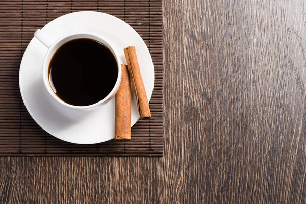 Cup of black coffee on table. Top view white cup and cinnamon sticks on porcelain saucer. Close up fresh and aromatic hot drink in cafe. Morning coffee break. Rustic wooden table with bamboo mat.