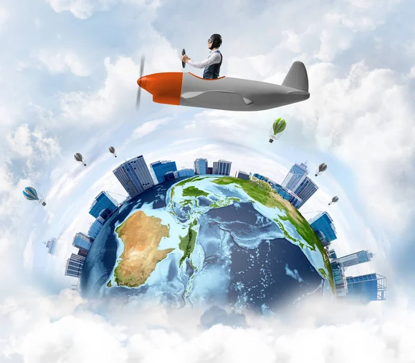 Young man in aviator hat with goggles driving propeller plane. Traveling around the world by airplane concept. Funny man flying in small airplane in sky with clouds. Round earth with skyscrapers.