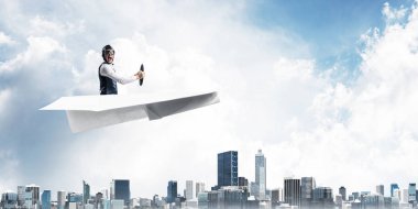 Kağıt uçakta oturan pilot ile iş motivasyonu kavramı. Havacı şapkalı komik adam ve şehrin üzerinde kağıt uçak süren gözlükler. Rüyada gökyüzünde uçmak. Yüksek binalarile modern megapolis.
