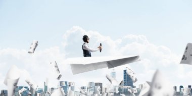 Havacı şapkalı işadamı ve gözlükşehir manzarası üzerinde kağıt uçak sürüyor. Mavi gökyüzünün arka planına bulutlar dökülen kağıtlar. İfade pilotu büyük kağıt uçak eğlenmek.