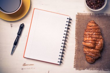 Açılmış not defteri ve bir fincan kahveyle ahşap masada çörek.