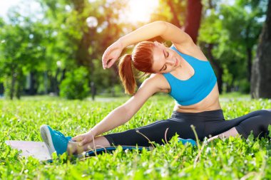 Güneşin doğuşunda spor salonunda esneme hareketleri yapan güzel bir kız. Aktif giyimli genç bir kadın yaz günü yeşil çimlerde oturuyor. Sabah sporu ve sağlıklı yaşam tarzı.