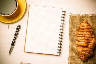 Açılmış not defteri ve bir fincan kahveyle ahşap masada çörek.