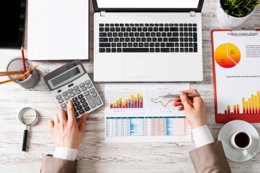 Ofis masasında mali analizcilerle çalışan bir iş adamı. İnsan eliyle veri hesaplayan üst düzey ofis işyeri. Dizüstü bilgisayarı, hesap makinesi, çizelgesi ve kahve fincanı olan düz bir masa..