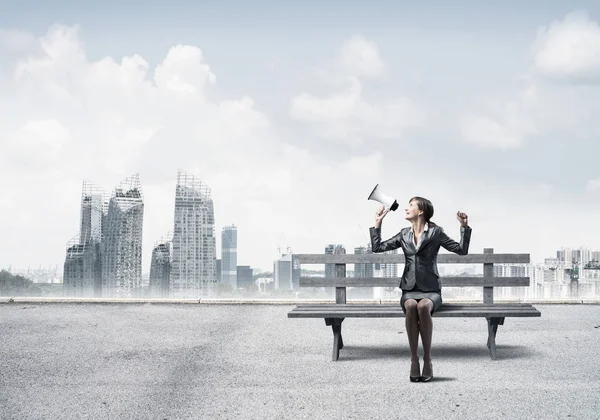 Business woman with megaphone sitting on wooden bench. Female speaker shouting in loudspeaker outdoors. Modern cityline panorama with cloudy sky. Business marketing and announcement.