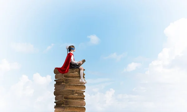空に古い本の山の上に座って読書マスクとケープの小さな子供 — ストック写真