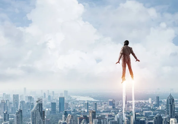 Businessman in suit and aviator hat flying in blue sky as superhero. Back view of corporate manager as superman launching upwards with jet flame above modern city. Successful business startup