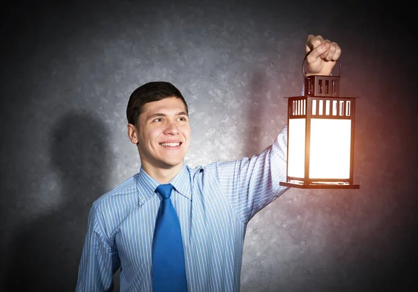 Happy businessman holding glowing lantern on background grunge wall. Front view of young man in shirt and tie looking for something in dark. Smiling business person lighting his way with lantern.