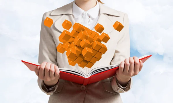 Young woman holding open book with flying 3d orange cubes. Hi-tech concept with virtual reality element. Closeup open book in female hands. Elegant lady in business suit on sky background.