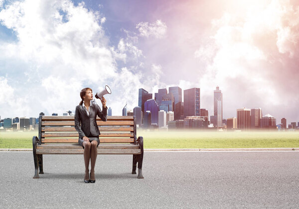 Business woman with megaphone sitting on wooden bench. Female speaker shouting in loudspeaker outdoors. Modern cityline panorama in sunny day. Business marketing and announcement.
