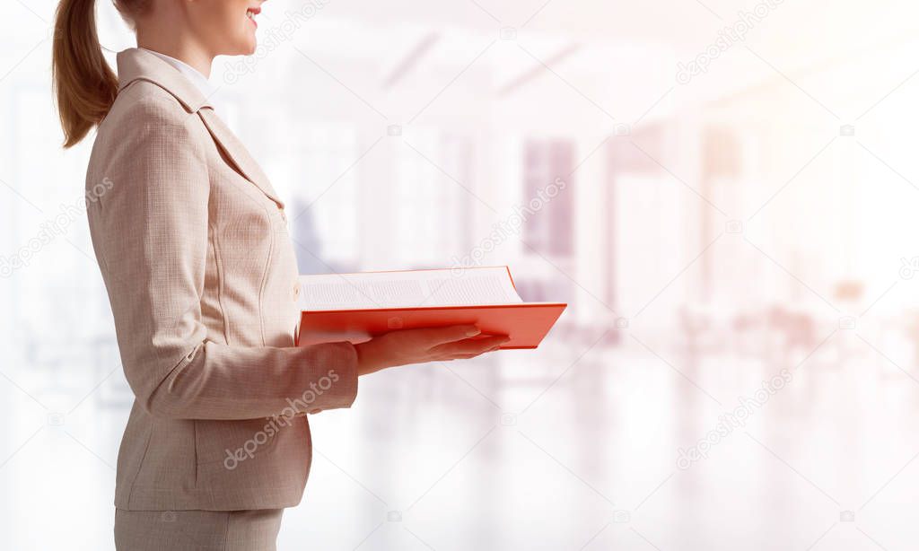 Businesswoman holding open notebook on background of blurred office interior. Training courses and workshops concept. Open book with red cover in female hands. Side view elegant lady in business suit.