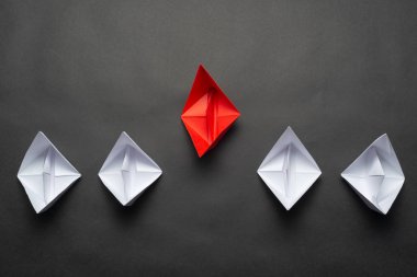 Siyah arka planda kağıt gemiler. Yaratıcı yenilik ve liderlik ticari konsepti. Beyaz teknelerin önünde düz kırmızı origami lideri yatıyordu. Şirket stratejisi ve başarılı çözüm.