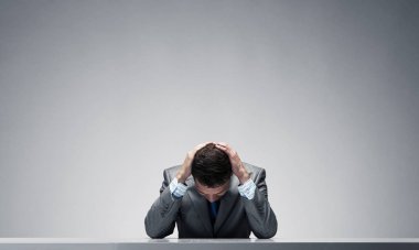 Elleri başının üstünde masada oturan sinirli bir adam. İşadamı iş yerindeki baskıdan dolayı stresli. İşsizlik ve kariyer sorunları. Mutsuz bir adamın portresi gri arka planda takım elbise ve kravat takıyor..