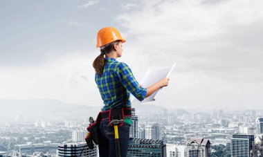 Teknik planla birlikte sabit şapkalı kadın mühendis. Kareli mavi gömlekli kadın mimarın şehrin arka planındaki inşaatı teftiş edişinin yan görüntüsü. Proje yönetimi inşa ediliyor