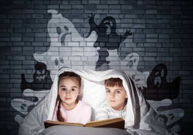 Sevimli kız ve erkek yatakta çocuk kitap okuma. El feneri olan çocuklar battaniyenin altında yatıyorlar. Pijamalı çocuklar ve komik hayaletler gri duvara geri siluetler. Sihirli peri masalları okuyan çocuklar