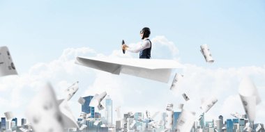 Havacı şapkalı işadamı ve gözlükşehir manzarası üzerinde kağıt uçak sürüyor. Mavi gökyüzünün arka planına bulutlar dökülen kağıtlar. İfade pilotu büyük kağıt uçak eğlenmek.