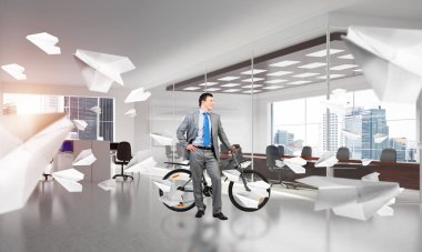 Kendinden emin ve başarılı bir adam iş elbisesi giyiyor ve bisikletiyle birlikte rahat çalışma alanında uçan kağıt uçaklarla kravat takıyor. İş adamı büyük pencereli modern ofiste bisikletle dinleniyor..