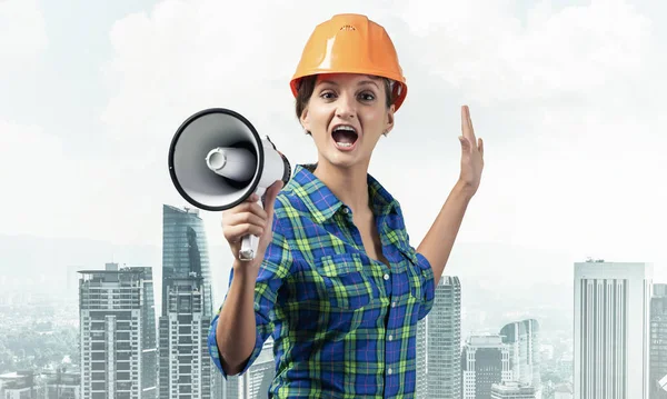 戴着安全帽的有表情的女人对着扩音器大喊 具有城市景观背景的张大嘴的年轻情感建筑工人的肖像 新闻广播和广告 — 图库照片