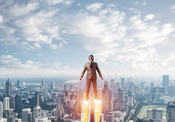 Businessman in suit and aviator hat flying in blue sky as superhero. Business person as superman launching upwards with jet flame above modern downtown. Successful business startup. Career growth